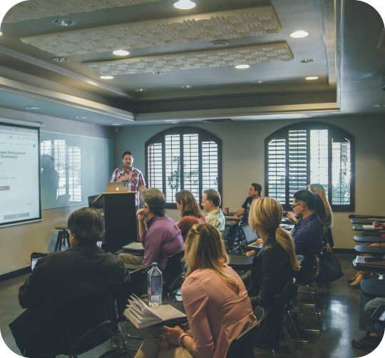 Ein Mann präsentiert lächelnd in einem Seminarraum, Zuhörer sind aufmerksam.