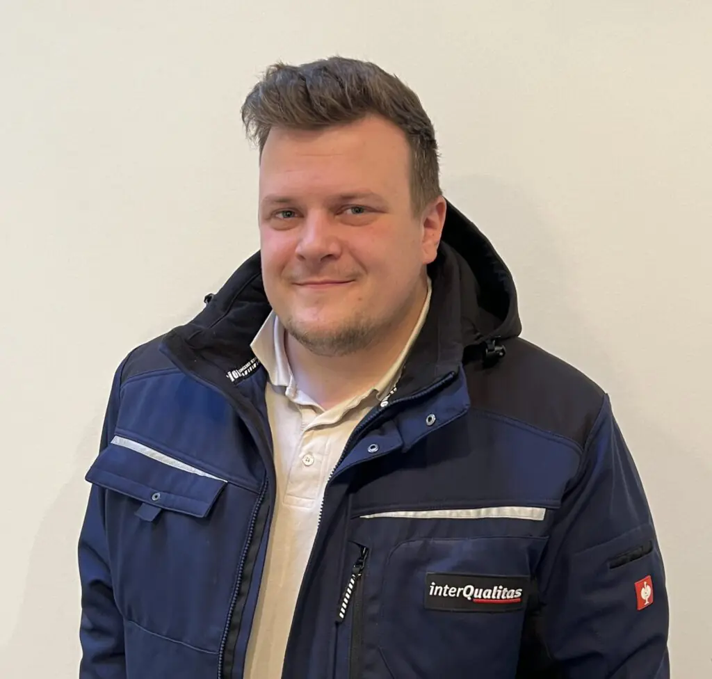 Mann lächelt leicht, trägt dunkelblaue Jacke mit "interQualitas"-Logo; kurzer, gestylter Haaransatz.