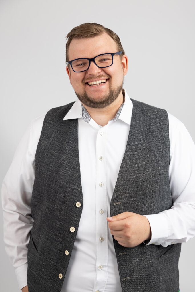 Ein lächelnder Mann mit Brille, weißem Hemd und grauer Weste vor grauem Hintergrund.